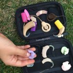 Description de la photo. Main d'enfant attrapant une empreinte d'oreille servant à fabriquer les embouts des appareils auditifs. D'autres empreintes, des appareils auditifs et des implants cochléaires sont contenus dans une boite noire posée sur l'herbe. Fin de la description de la photo.