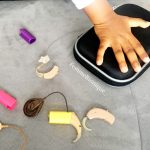 Description de la photo. Une main d'enfant posée sur une boite noire d'un implant cochléaire. A côté se trouve 2 vieux appareils auditif, 2 implants et des caches d'implant colorés. Fin de la description de la photo.