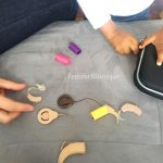 Description de la photo. Un enfant s'amuse à ouvrir et fermer une boite noire d'un implant cochléaire. A côté se trouve 2 vieux appareils auditif, 2 implants et des caches d'implant colorés. Une main d'adulte s'apprête à attraper un appareil auditif. Fin de la description de la photo.